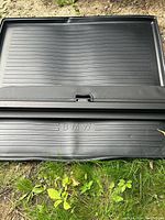 Black BMW trunk cover laying flat on grass, showing embossed BMW logo and horizontal textured lines, slight surface warping visible