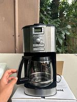 Front view of Cuisine Art programmable coffee maker showing digital display, program buttons, and glass carafe held by hand.