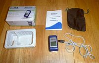 Pulse oximeter with user manual, original box, black soft pouch, white braided lanyard on wooden table