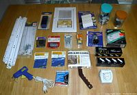 Photo showing various hardware and household items including jars with small parts, curtain hardware, electrical items, and tools arranged on a wooden surface.