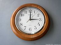 Front view of wooden wall clock with metal ring, white face, black numerals and hands showing 3:00.
