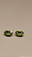 Pair of oval-cut apple green Peridot gemstones on beige background, showing their color and clarity.