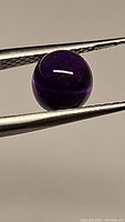 Close-up photo of the deep purple round cabochon amethyst stone held in tweezers showing its smooth polished rounded surface and natural inclusions visible inside the gemstone.