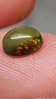 Close-up photo of an 8x6mm oval cabochon Ethiopian black opal gemstone showing bright flashes of red, green, and orange colors against a mostly charcoal background.