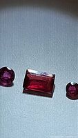 Three natural rubies displayed under light, showing an emerald cut rectangular ruby centered between two round cut rubies with bright deep red to pink hues.
