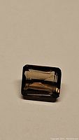 Single natural smokey quartz gemstone, emerald cut, placed on a plain surface for a clear view of its cuts and clarity. The stone has a reflective surface showing its faceting.