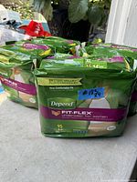 Photo of several green packages of Depend Fit-Flex underwear for women, size XL on a concrete surface with plants in the background.