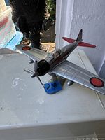 A vintage tin toy airplane on a stand with red circular wing markings and black propeller. The pull string mechanism is visible on the back of the stand.