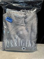 Grey hoodie folded inside clear plastic packaging with label #3a, showing white text "ORRIGINA" and maple leaf logo.