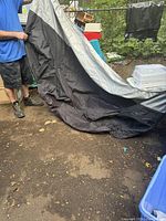 Photo showing a person holding a large black and silver motorcycle cover outdoors, with the cover spread out partly on the ground.