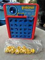 Photo of red Perfection game board with blue grid and yellow puzzle pieces in plastic bag placed in front of board.