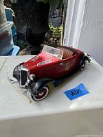 Front side view of red and black die cast 1934 Ford Roadster model car with Coca-Cola logos on hood and side, showing whitewall tires and chrome grille.