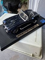 Front-side angled view of dark blue die cast Shelby Cobra model on a black display base labeled with model name.
