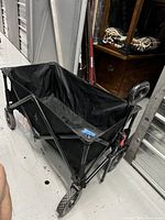 Black folding wagon fully assembled, showing fabric and metal frame on floor next to storage doors.
