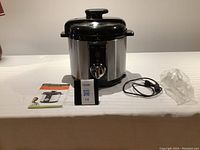 Full view of the Wolfgang Puck electric pressure cooker with power cord, manual, and accessory on a white surface.