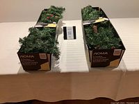 Two boxes containing NOMA 9-foot Carolina pre-lit garlands placed side by side on a table, showing the packaging and partial garland details such as greenery, pine cones, and red berries.