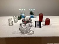 Full view of all items on white tablecloth including crystal Santa sleigh, two glass candle holders, two red flameless candles, glass bowl with candle and decorative fillers, and a pair of blue glass lanterns.