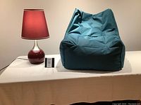 Photo showing a turquoise bean bag chair next to a lamp with a burgundy shade and ceramic base on a white surface.