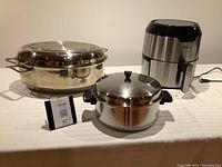 Tfal electric fryer, stainless steel roasting pan with grill, and large cooking pot with lid displayed side by side on white cloth-covered surface.