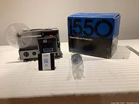 Full view of BerkeyKeystone 1550Z movie projector with clear plastic reel, original blue and black box, and accessories on table.