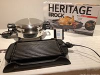 Wolfgang Puck electric grill/griddle, stainless steel skillet with lid, and boxed Heritage Rock roasting pan shown together on table.