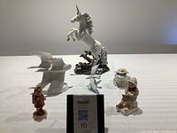 Front view of six porcelain and ceramic figurines on a white surface, including a large unicorn, white geese, seagull, frog, and two Goebel Hummel figurines.