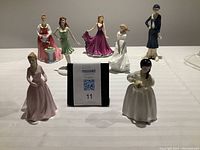 Seven porcelain figurines displayed on a white tablecloth, including various female figures in different colored dresses and poses.