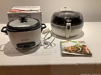 Black & Decker rice cooker and steamer next to T-Fal Actifry and recipe booklet on table.