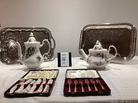 Two Royal Albert fine china teapots, two silver serving trays with floral etching, and two sets of sterling silver cake forks in cases arranged on a table.