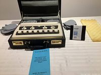 Closed briefcase opened to show vintage TENS/EMS control unit with white dials, electrode pads, and blue manual on white surface.