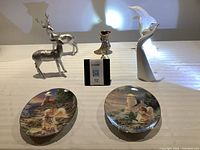 Full view of two Angels Light collection wall plates, silver plated stag and doe statues, colorful vintage ladies boot and bell figurines arranged on table under light.