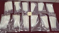 12 sealed packages of white plastic zip ties arranged in two rows on a red surface. Each package contains 100 zip ties.