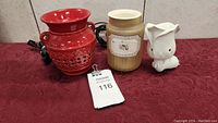 Three Scentsy warmers on a red fabric surface with power cords visible. Warmers include a red ceramic open lattice design, beige jar style with label, and white bunny shape.