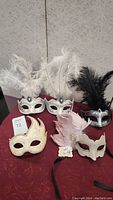 Five masquerade masks on burgundy fabric with feathers and decorative accents.