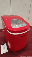 Red Frigidaire countertop ice maker with triangular transparent lid and front logo visible, placed on red surface.
