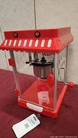Photo of side and front showing red popcorn machine with metal kettle and crank handle inside clear panels.