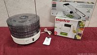 K-Tel food dehydrator with clear trays and power cord on red surface, beside boxed OXO Good Grips and Starfrit slicers.