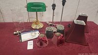 Photo showing full lot on red cloth background including green banker's lamp, pink fabric lampshades, metal bases, brown lampshades, lamp sockets and wiring.
