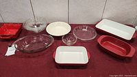 Full view of all bake and oven ware dishes laid out on a red surface against white tiled wall.