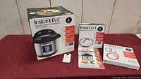 Photo of boxed new Instant Pot Ultra 10-in-1 multi-use programmable pressure cooker with accessories including glass lid box, cookbooks and sealing rings on red surface against tiled wall.
