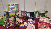 Various toys and games on red table including Crayola Widescreen Light Designer, Play-Doh, toy vehicles, figurines, puzzles and art supplies