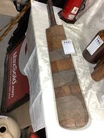 Wooden cricket paddle on display on a patterned tablecloth with auction labels, showing full length and handle.