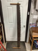 Full view of the long antique crosscut saw hanging vertically on a white door. Shows wood handles on both ends and the full length of the saw blade with teeth.