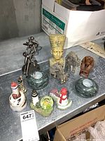 Photo showing assortment of collectible figurines including lighthouse figures, marble candle holders, metal sculpture, resin elephants, and pharaoh busts arranged on a surface.