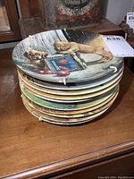 Photo showing a stack of 10 round collector plates with dog illustrations, one plate on top showing two golden retriever puppies near a box and fruit.