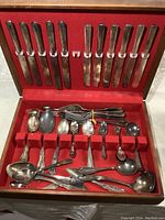 Top-down view of the open wooden chest displaying twelve butter knives in the lid, slots filled with an assortment of spoons, forks, and ladles on red felt lining.
