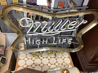 Front view showing illuminated Miller High Life lettering in white and yellow neon tubes within brass frame.