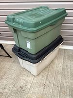 Stacked view of three Rubbermaid totes in green, black, and white colors against siding, showing size and wear.