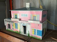 Front view of the two-story metal doll house with pink upper floor, white lower floor, painted windows, doors, and floral landscaping.
