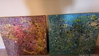 Pair of abstract paintings displayed side by side on the floor against a wall, showing one with pink, yellow, gray tones and the other with blue, green, white splatters on 30x30 canvases.